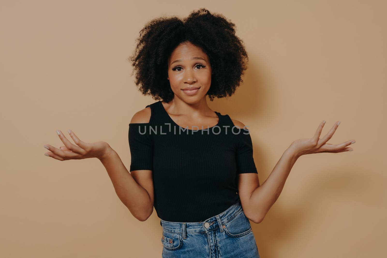 What should I do now. Confused unaware Afro woman with wavy curly hair, raises hands in bewilderment, looking confused, cannot make decision, isolated over beige studio background with copy space