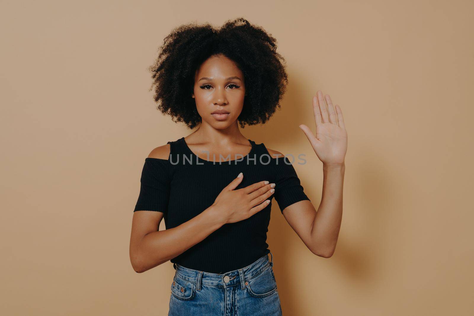 Dark skinned woman wearing casual shirt and jeans standing over isolated beige background, making swearing pose with hand on chest and fingers up, doing loyalty promise oath. Patriotism concept