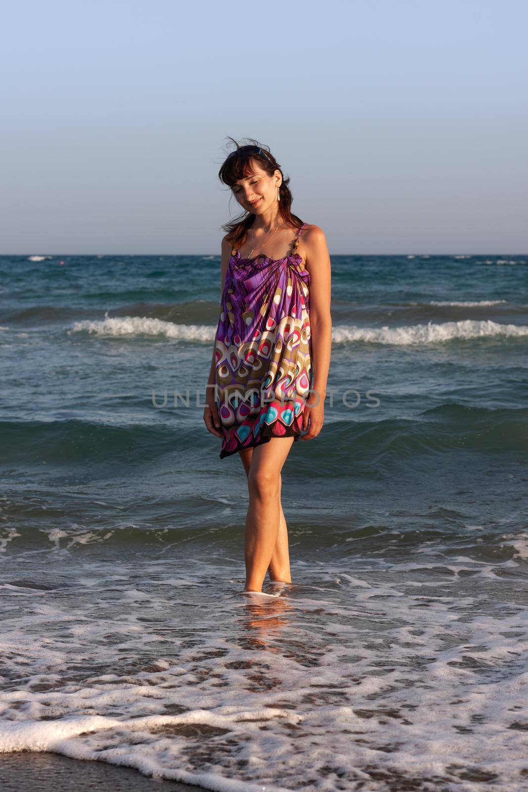 Young beautiful woman in a summer dress stands in the sea water on the sandy shore and enjoys the waves and the sea breeze