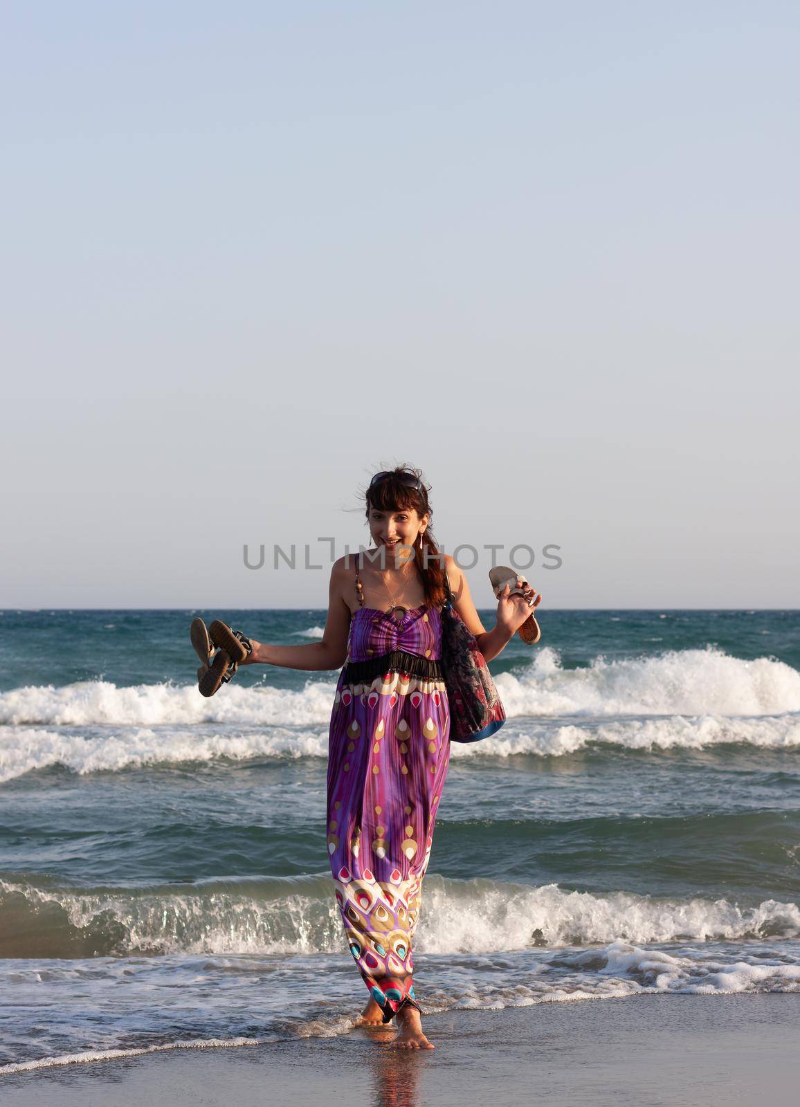 Young beautiful woman in a summer dress stands in the sea water on the sandy shore and enjoys the waves and the sea breeze