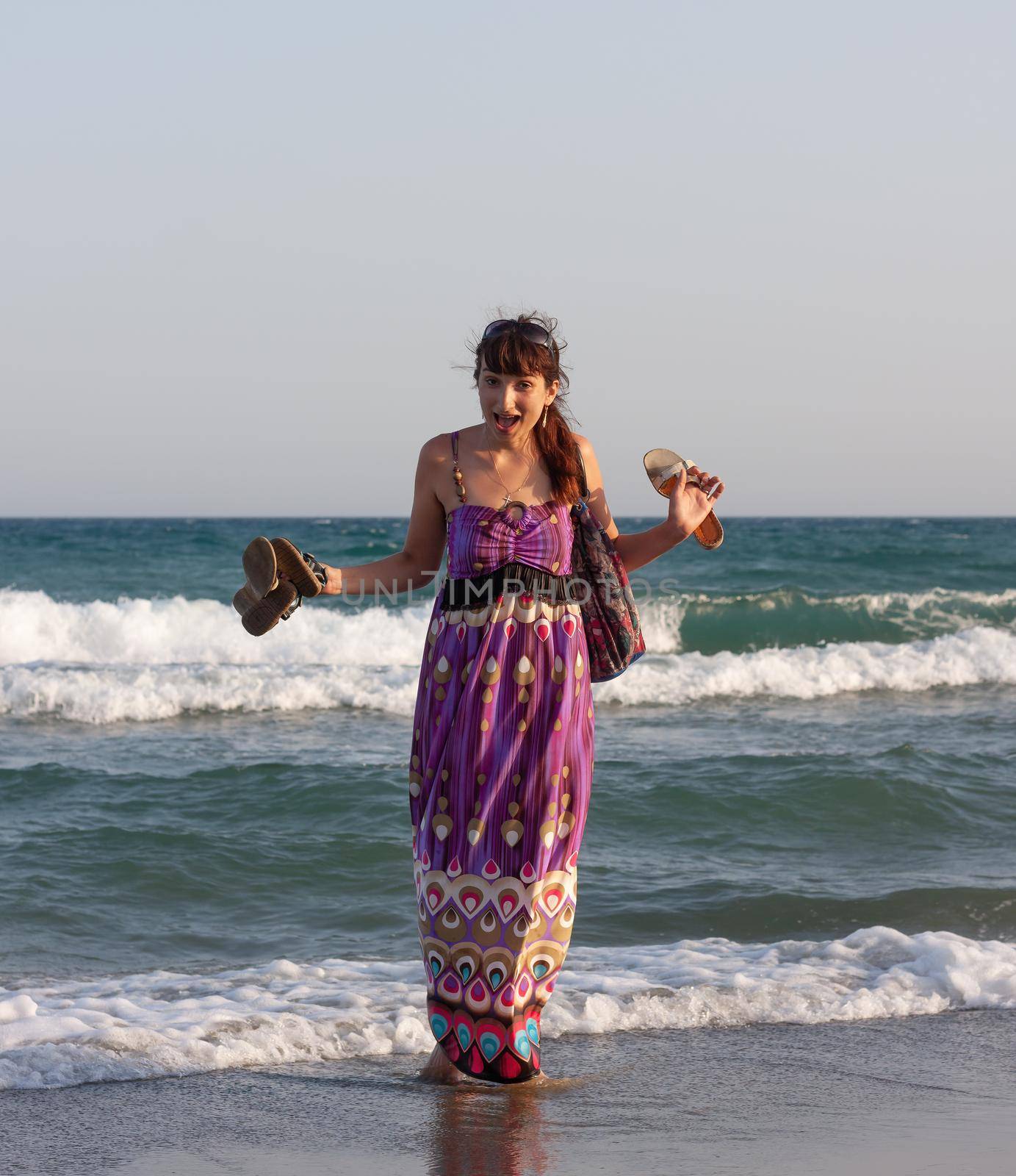 Young beautiful woman in a summer dress stands in the sea water on the sandy shore and enjoys the waves and the sea breeze