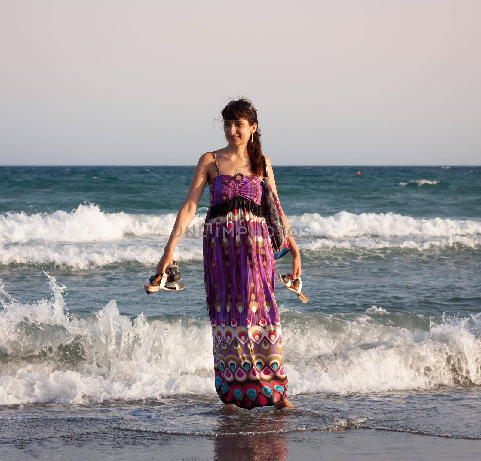 Young beautiful woman in a summer dress stands in the sea water on the sandy shore and enjoys the waves and the sea breeze
