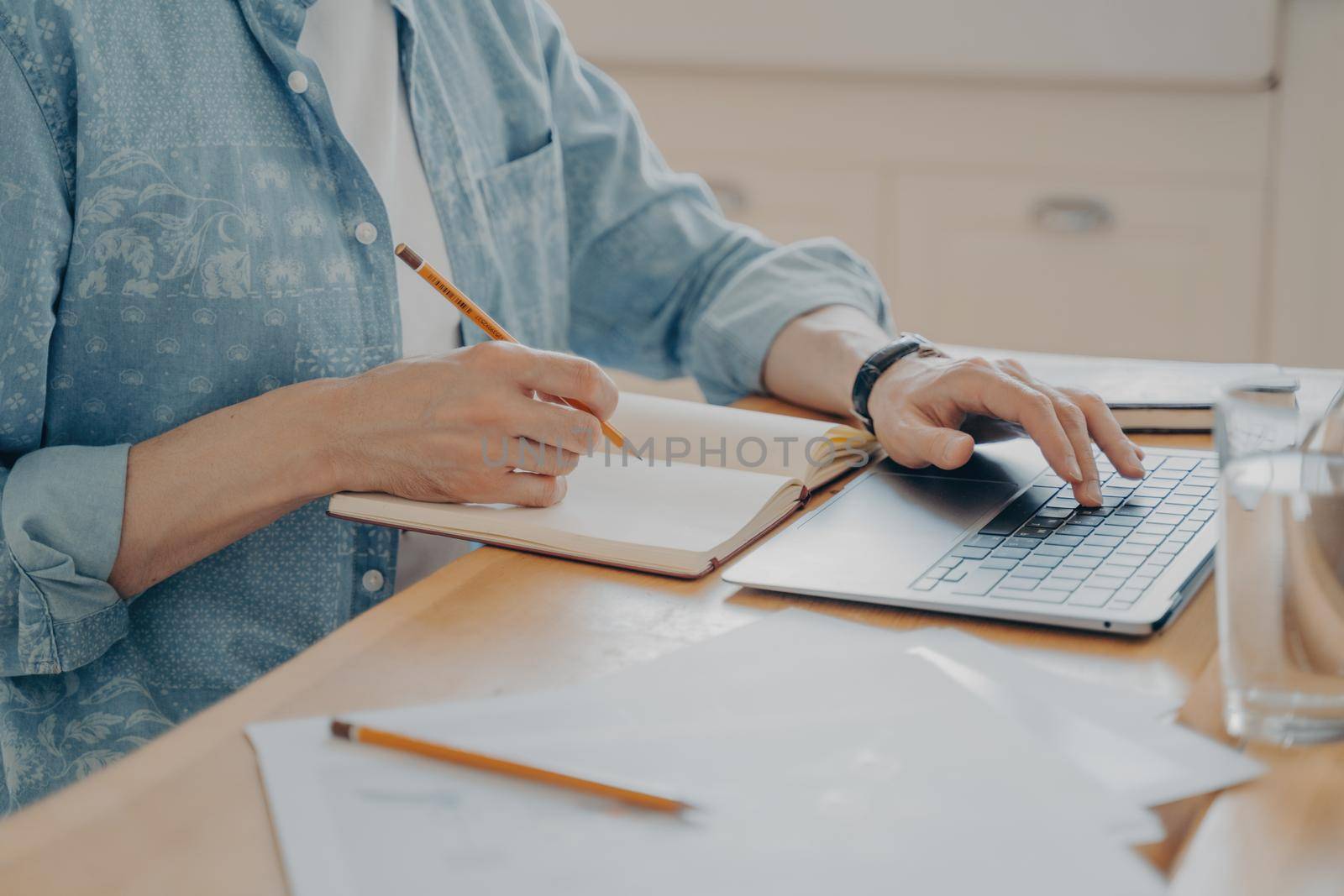 Close up picture of male hands typing on laptop for writing new article in website and making notes in agenda at kitchen wooden table. Concept of freelance and remote work from home