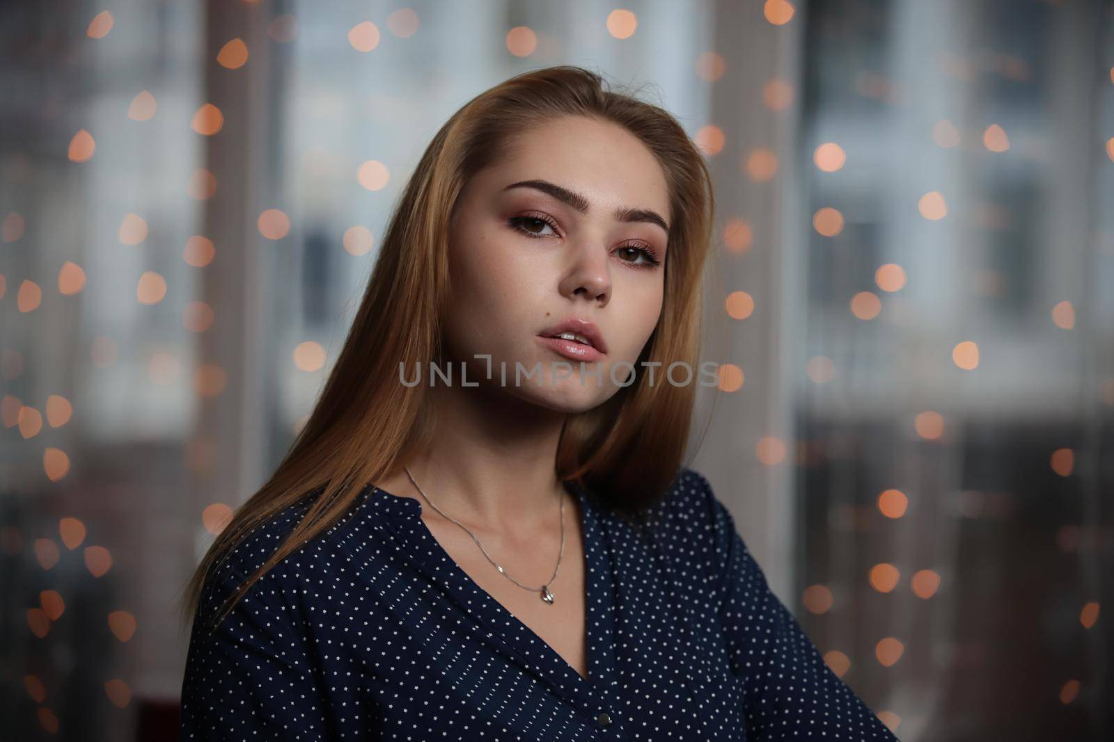 Portrait of beautiful young woman sitting near the window looking at camera over lights in background.Portrait