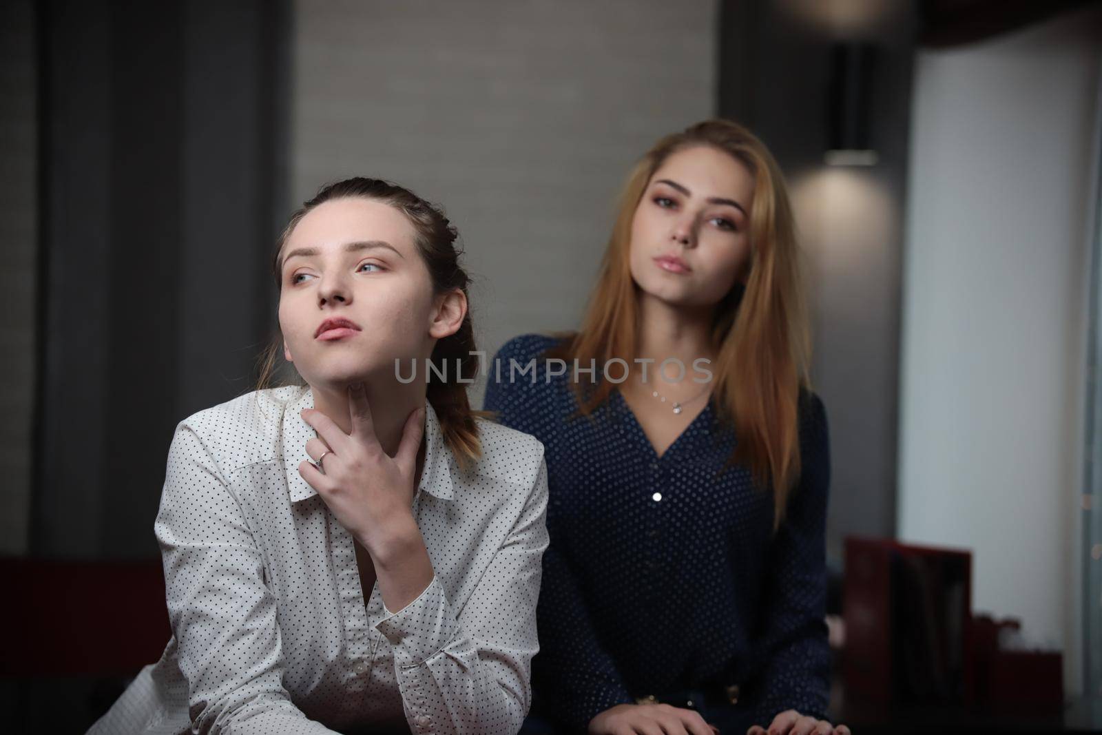 Portrait of two beautiful young women sitting at the table looking at camera over lights in background. Portrait