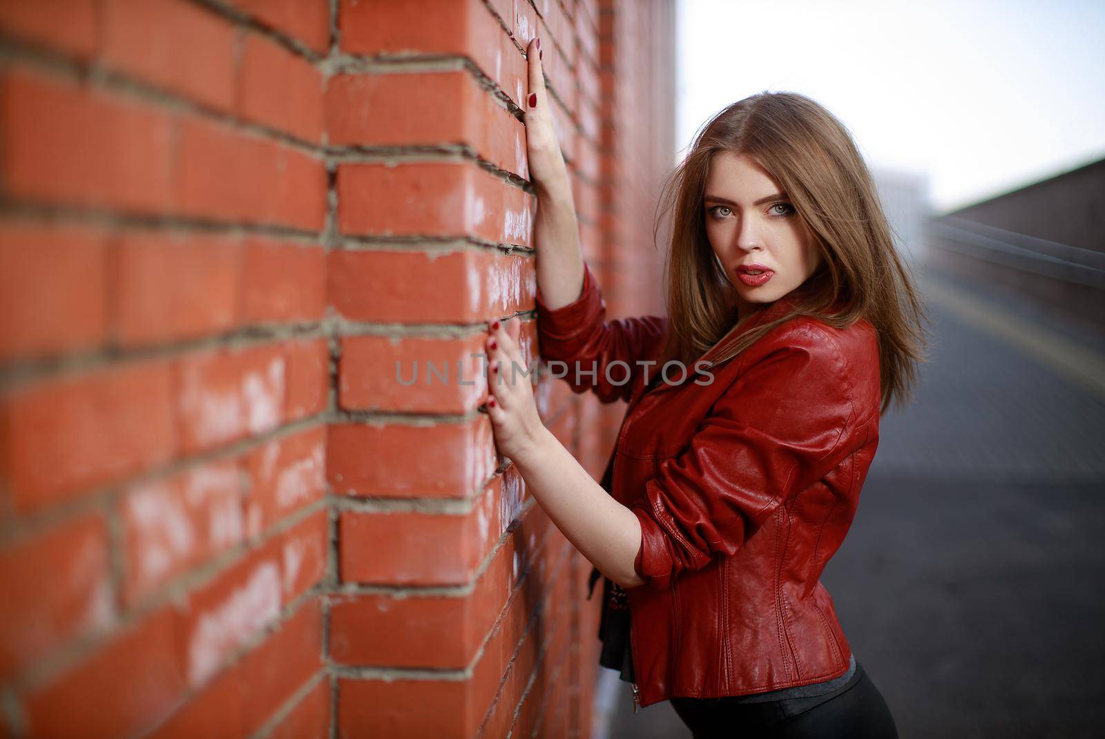Portrait of young woman in black leggings and red leather jacket standing near red brick wall