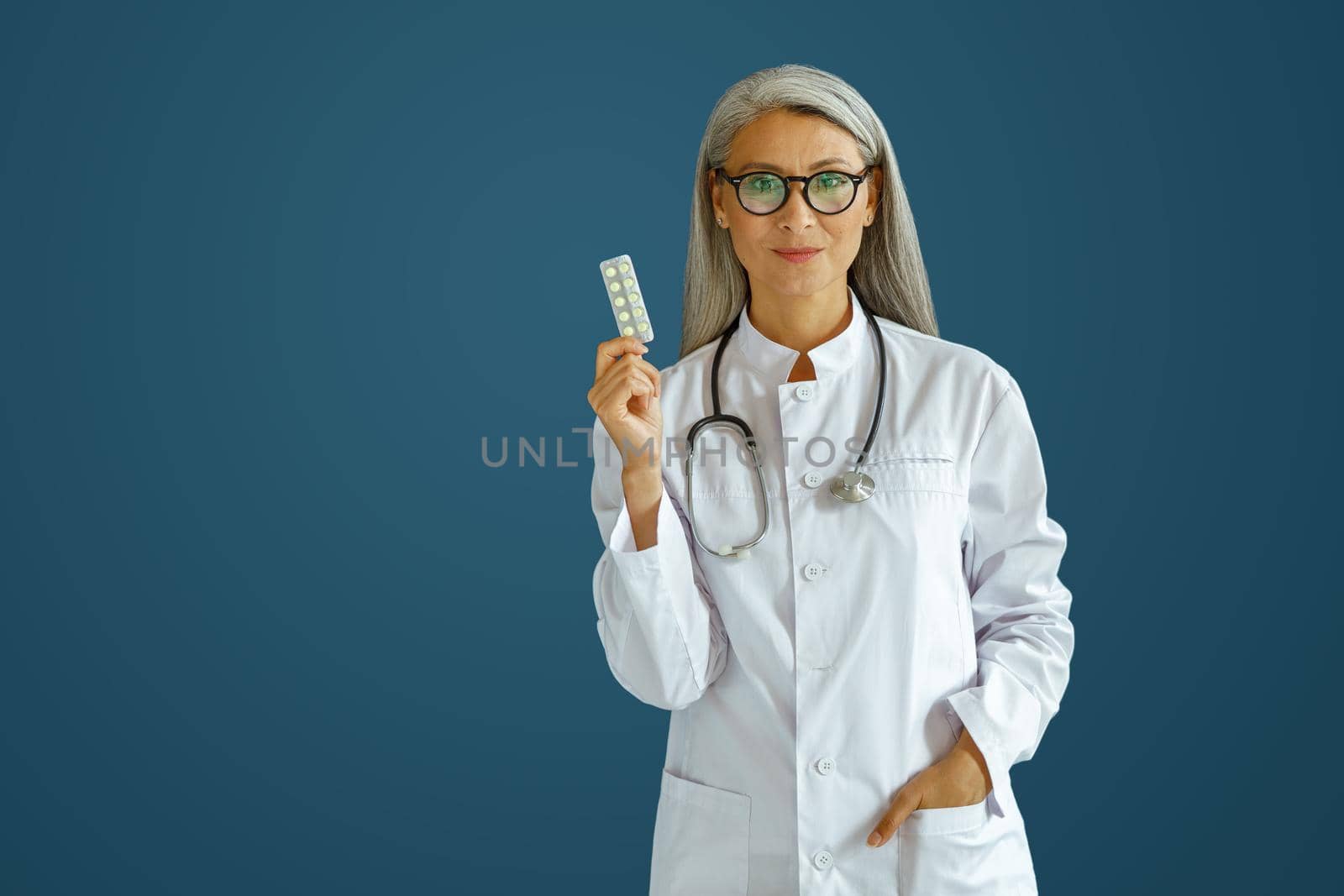 Positive mature woman therapist in white uniform shows pills blister standing on blue background in studio, space for text. Professional medical staff