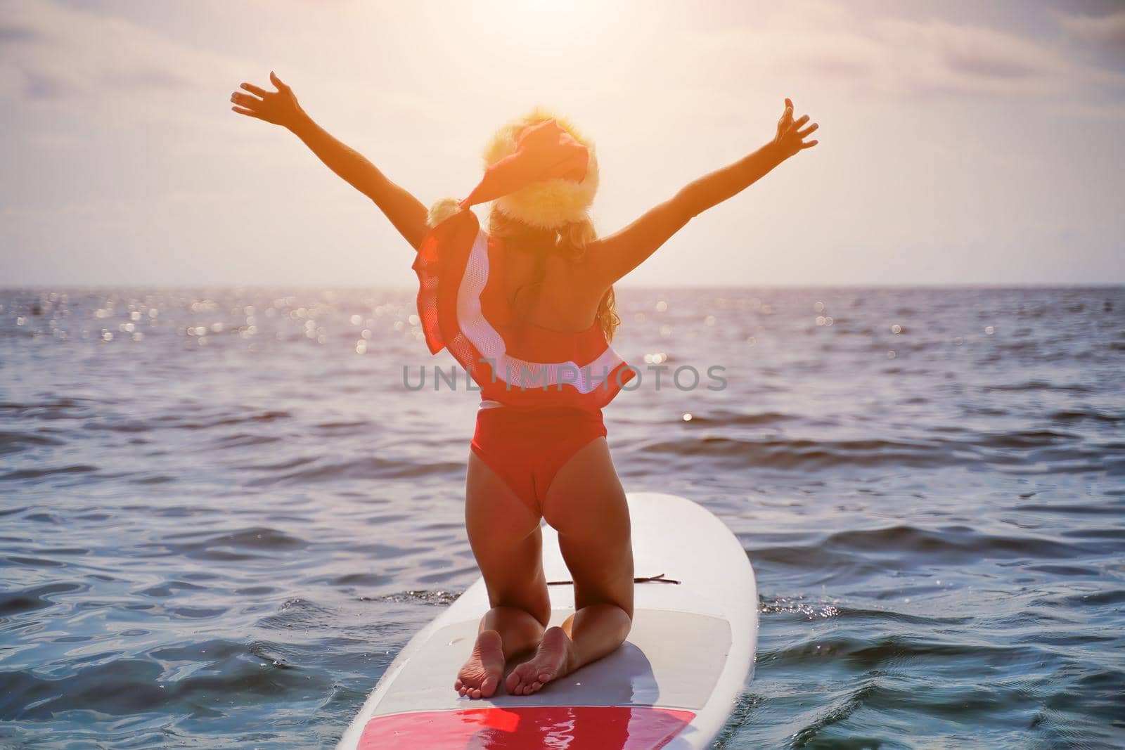 Happy young attractive brunette woman in red swimsuit and Santa hat, swimming on sup board in the sea. Christmas holiday vacation and travel concept. by panophotograph