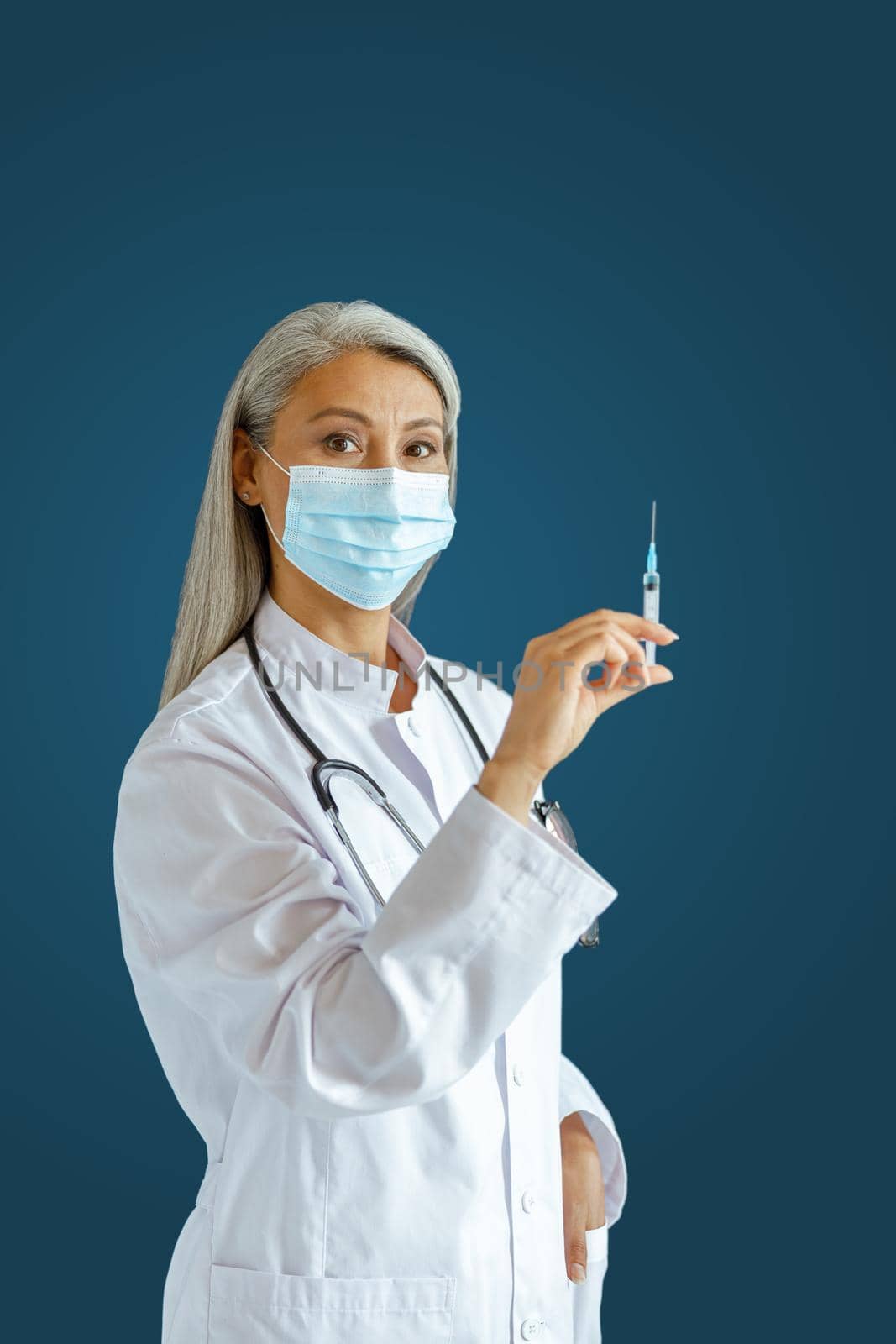 Silver haired Asian female therapist in uniform with protective mask holds syringe with medicine standing on blue background in studio. Vaccination day