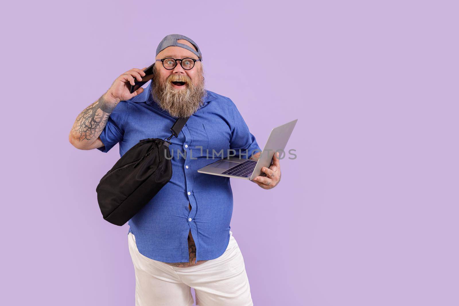 Funny bearded fat male person with laptop and crossbody bag talks on mobile phone standing on purple background in studio