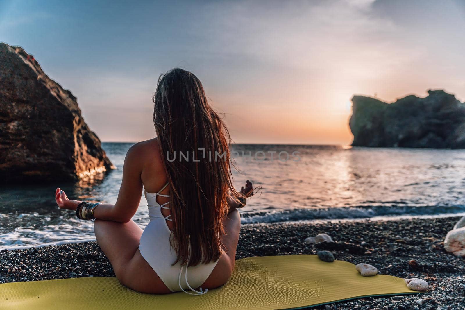 Young woman with long hair in white swimsuit and boho style braclets practicing outdoors on yoga mat by the sea on a sunset. Women's yoga fitness routine. Healthy lifestyle, harmony and meditation by panophotograph