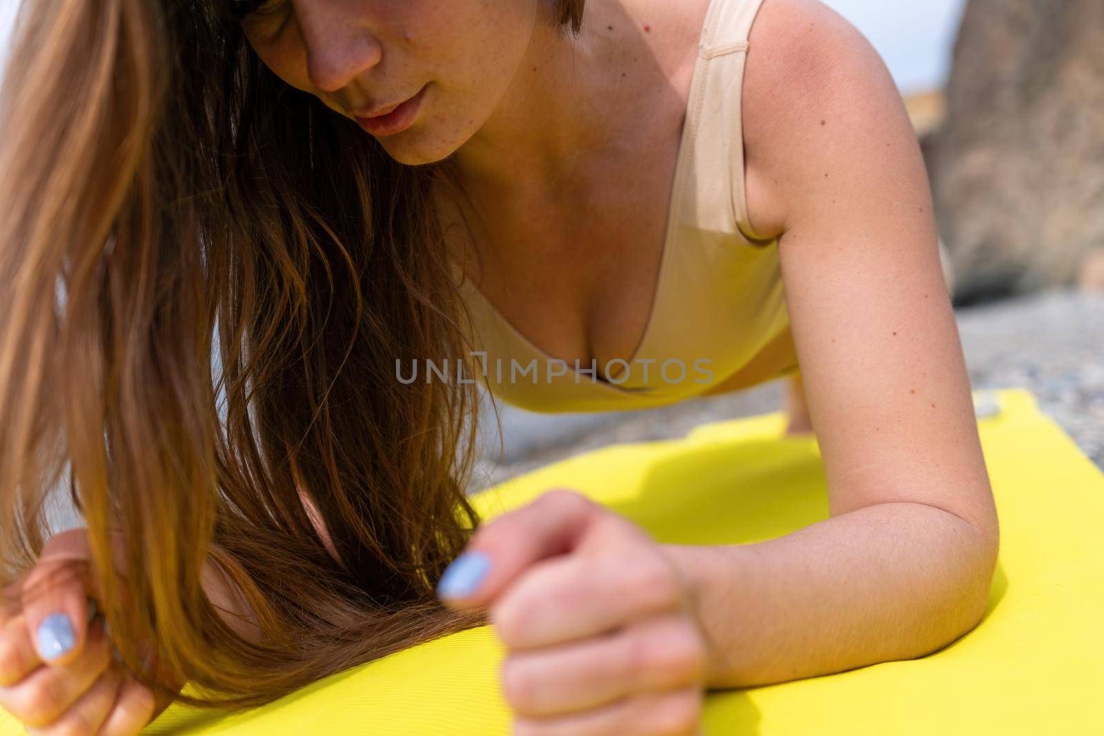 Young woman in swimsuit with long hair practicing stretching outdoors on yoga mat by the sea on a sunny day. Women's yoga fitness pilates routine. Healthy lifestyle, harmony and meditation concept.