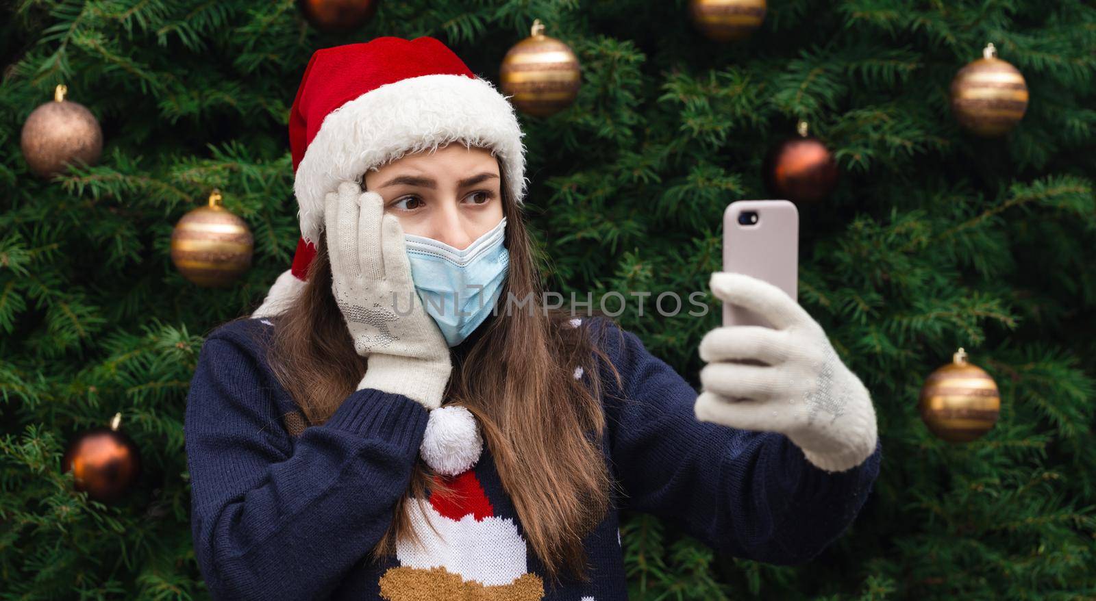 Christmas online greetings. Close up Portrait of woman wearing a santa claus hat and medical mask with emotion. Against the background of a Christmas tree. Coronavirus pandemic by lunarts