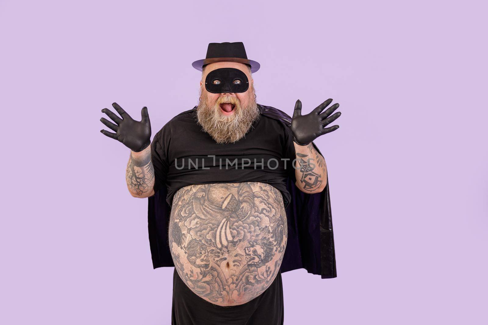 Excited mature male person with overweight wearing hero suit with cape and mask throws up hands standing on purple background in studio