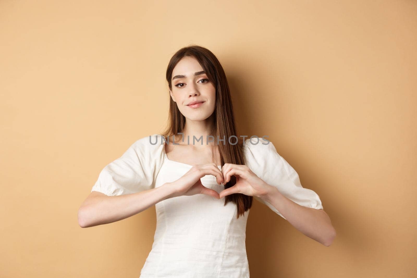 Valentines day and romance concept. Romantic caucasian girl in white dress showing heart gesture and smiling, say I love you, standing over beige background by Benzoix