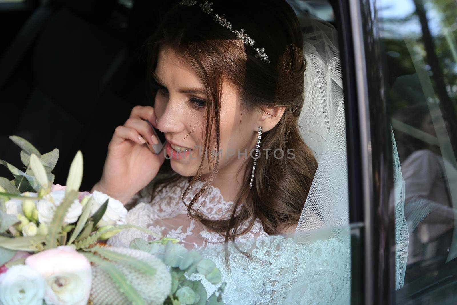 Wedding photo of the bride who is sitting in the car with a bouquet of flowers and talking on the phone.