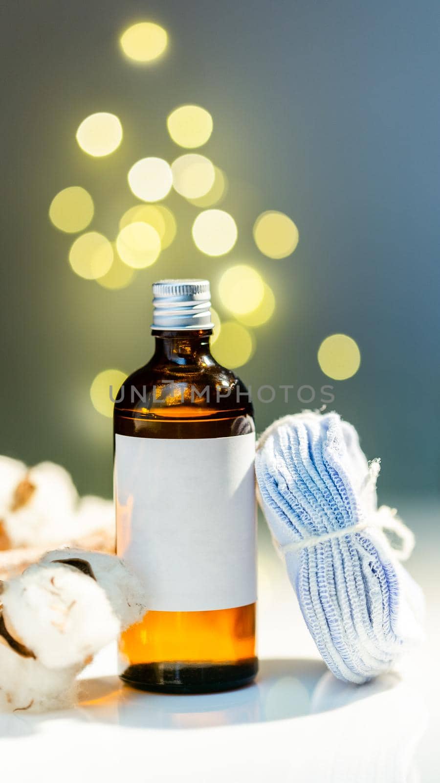 Festive face care product set. Toner in glass bottle and stack of reusable cotton round pads. White label mock up. Bokeh lights sparkling blur efect background
