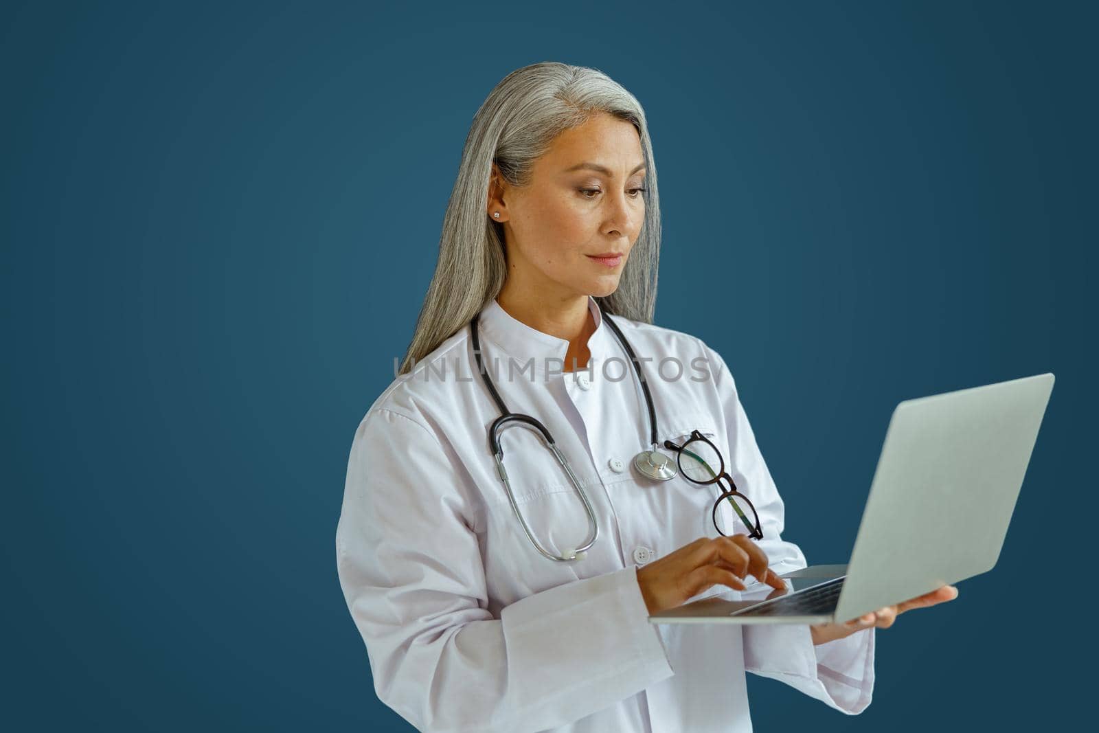 Concentrated grey haired woman doctor with stethoscope works on laptop standing on blue background in studio. Telemedicine patient service