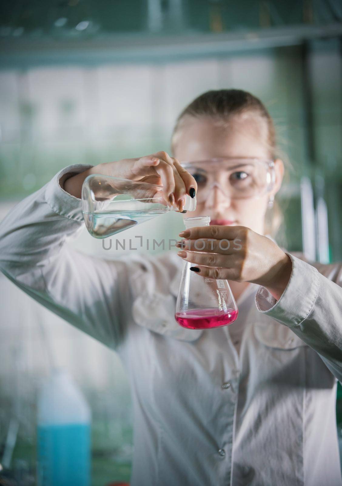Chemical laboratory. Young blonde woman holding two flasks with liquid in it. About to transfuse the liquid. Portrait