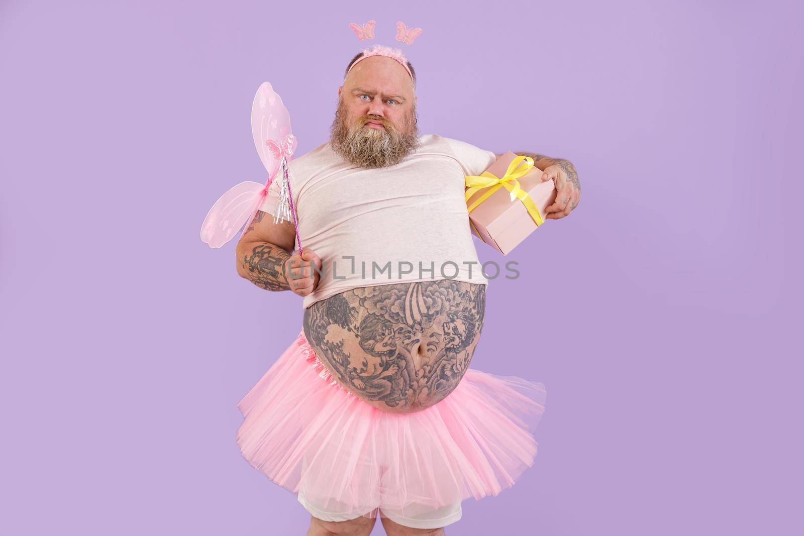 Funny bearded plump man in fairy costume with gift box grimaces on purple background by Yaroslav_astakhov