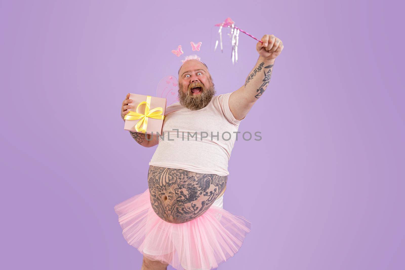 Excited man with overweight in fairy costume holds magic stick and present on purple background by Yaroslav_astakhov