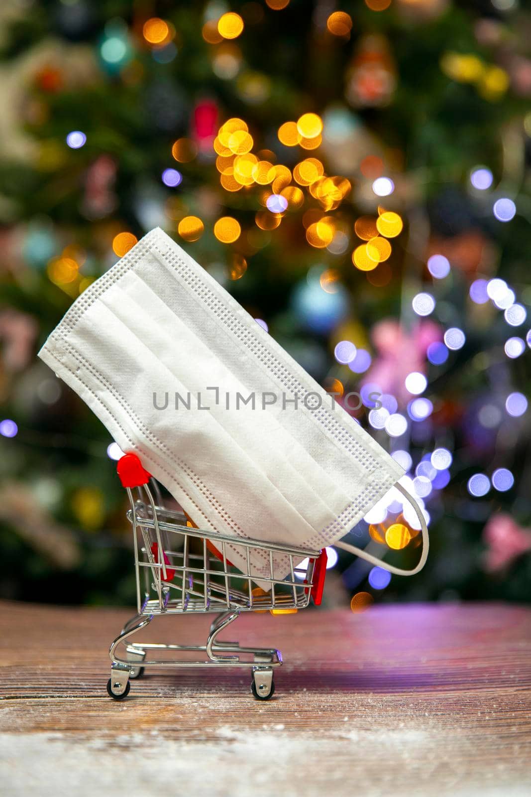 Protective safety mask in iron shopping basket near Christmas tree, Covid-19, coronavirus, holiday concept with copy space and bokeh lights background by Annebel146