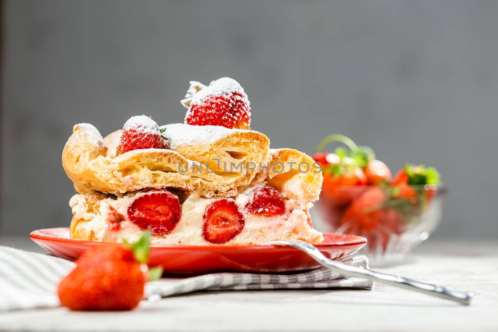 A piece of strawberry cake on a red plate iced with sugar powder over grey background