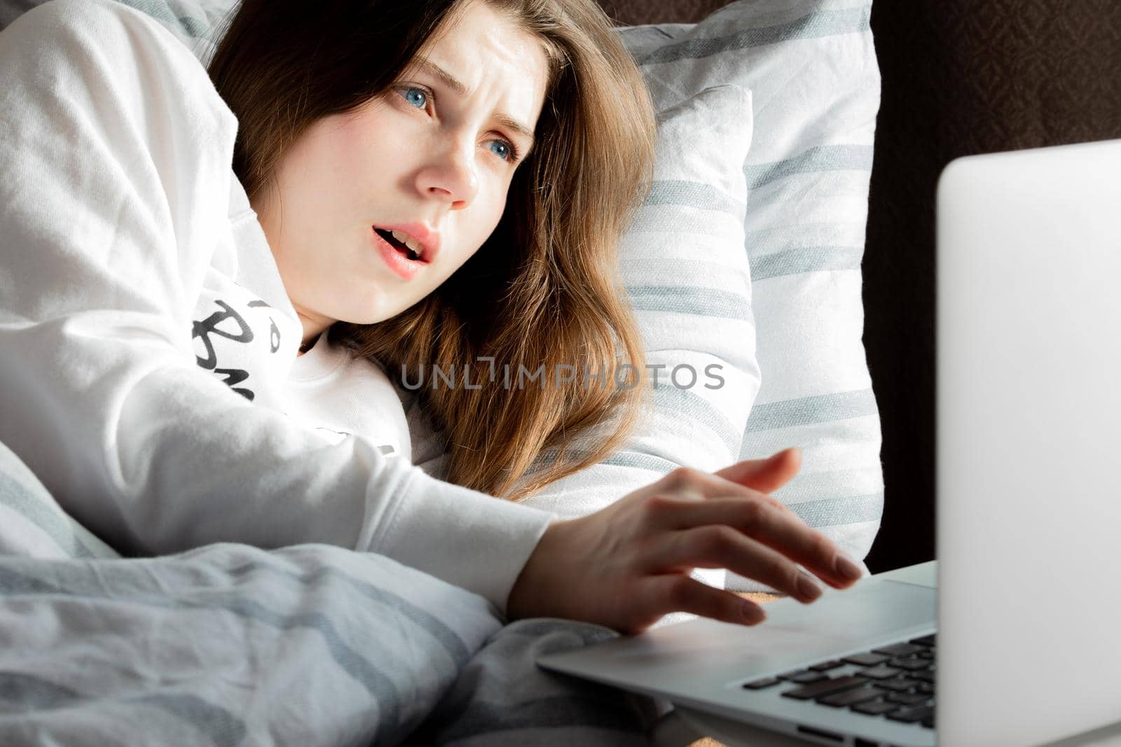 A young cute girl on a sunny day in a white jacket lying on the bed and looking at the laptop in surprise by lunarts