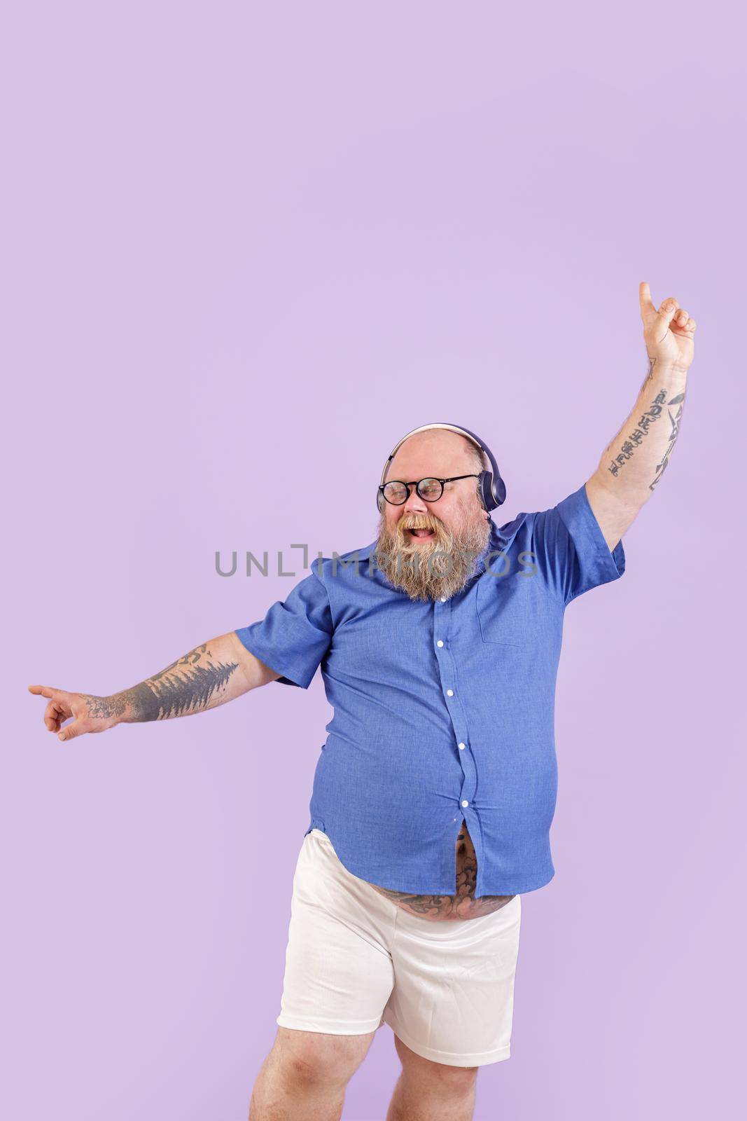 Positive mature bearded man with overweight in tight shirt and shorts enjoys favourite music with headphones dancing on purple background in studio