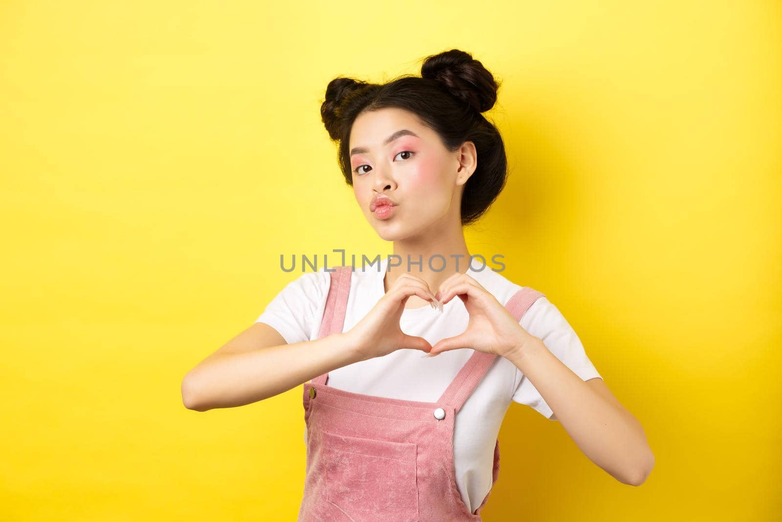 Silly japanese girl showing heart gesture and wishing happy Valentines day, pucker lips to kiss lover, standing on yellow background by Benzoix