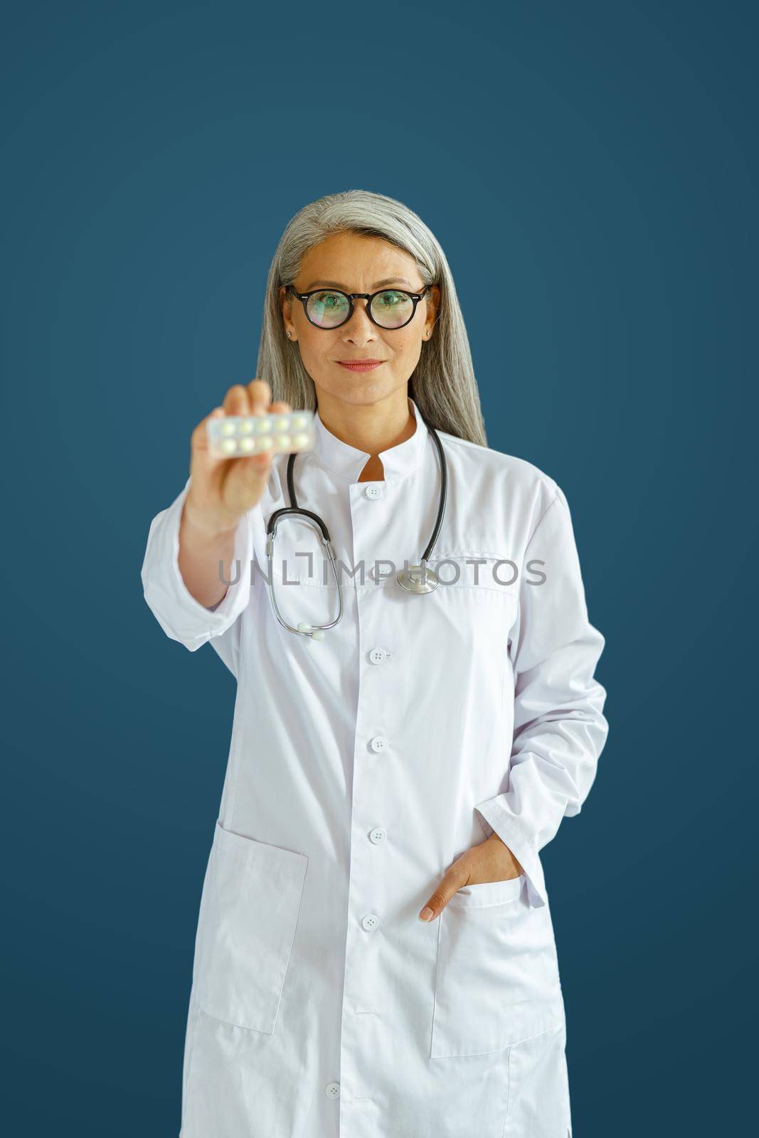Cheerful mature woman doctor in white robe shows pills blister posing on blue background in studio. Professional medical staff