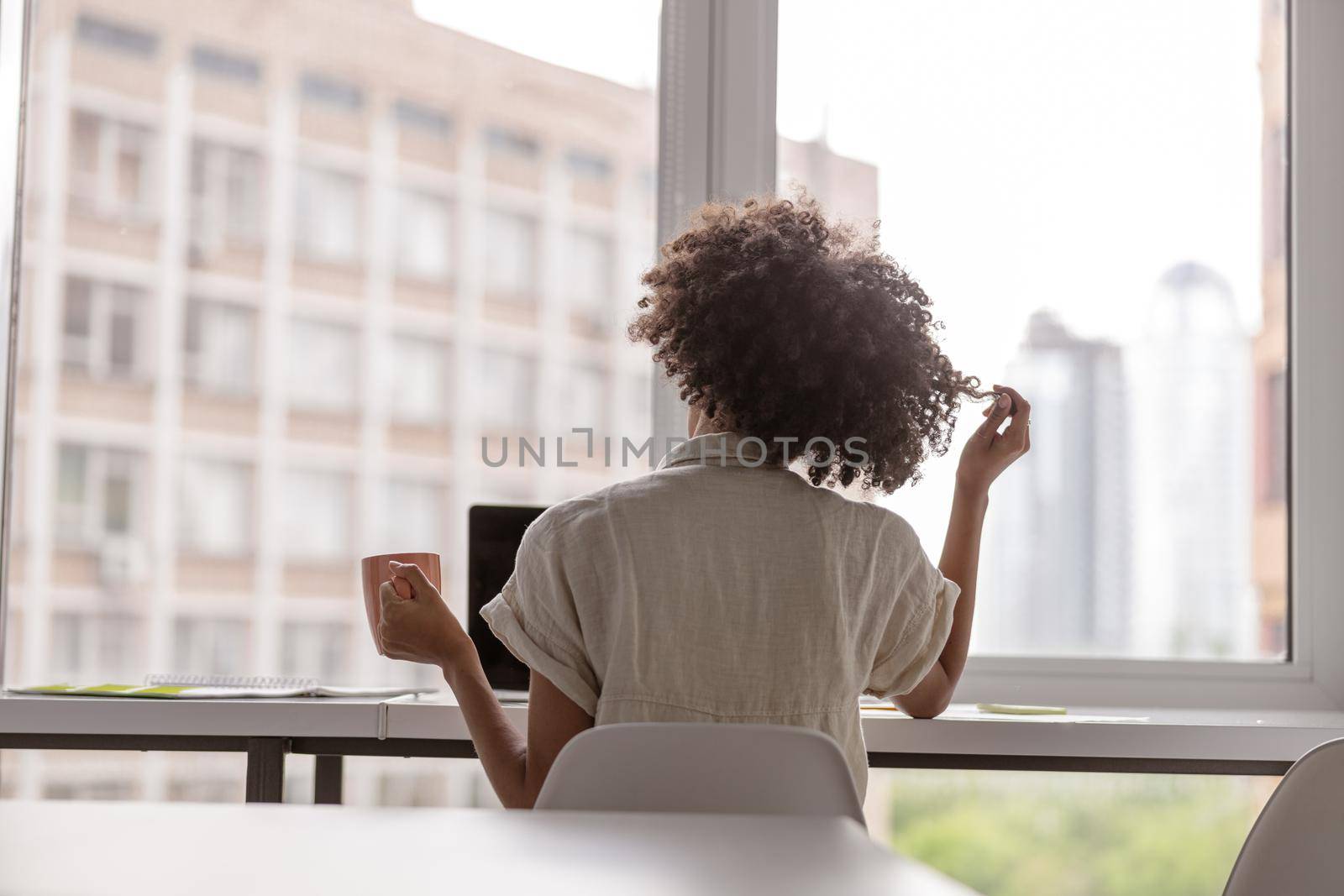 Back view of young woman sitting at desk near window while using laptop and holding cup. Copy space