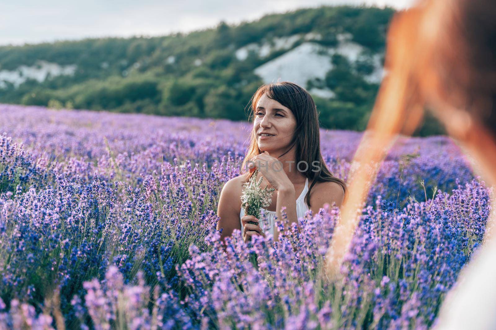 Close up portrait of happy young brunette woman in white dress on blooming fragrant lavender fields with endless rows. Warm sunset light. Bushes of lavender purple aromatic flowers on lavender fields. by panophotograph