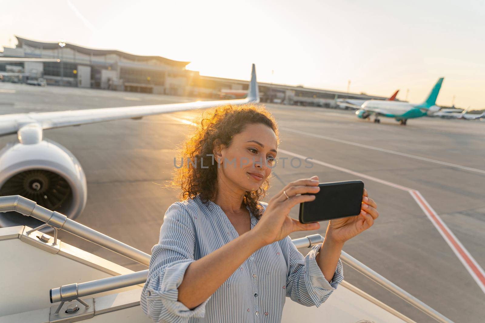 Smiling pretty lady enjoying her trip while making photo on her smartphone near plane. Travel concept