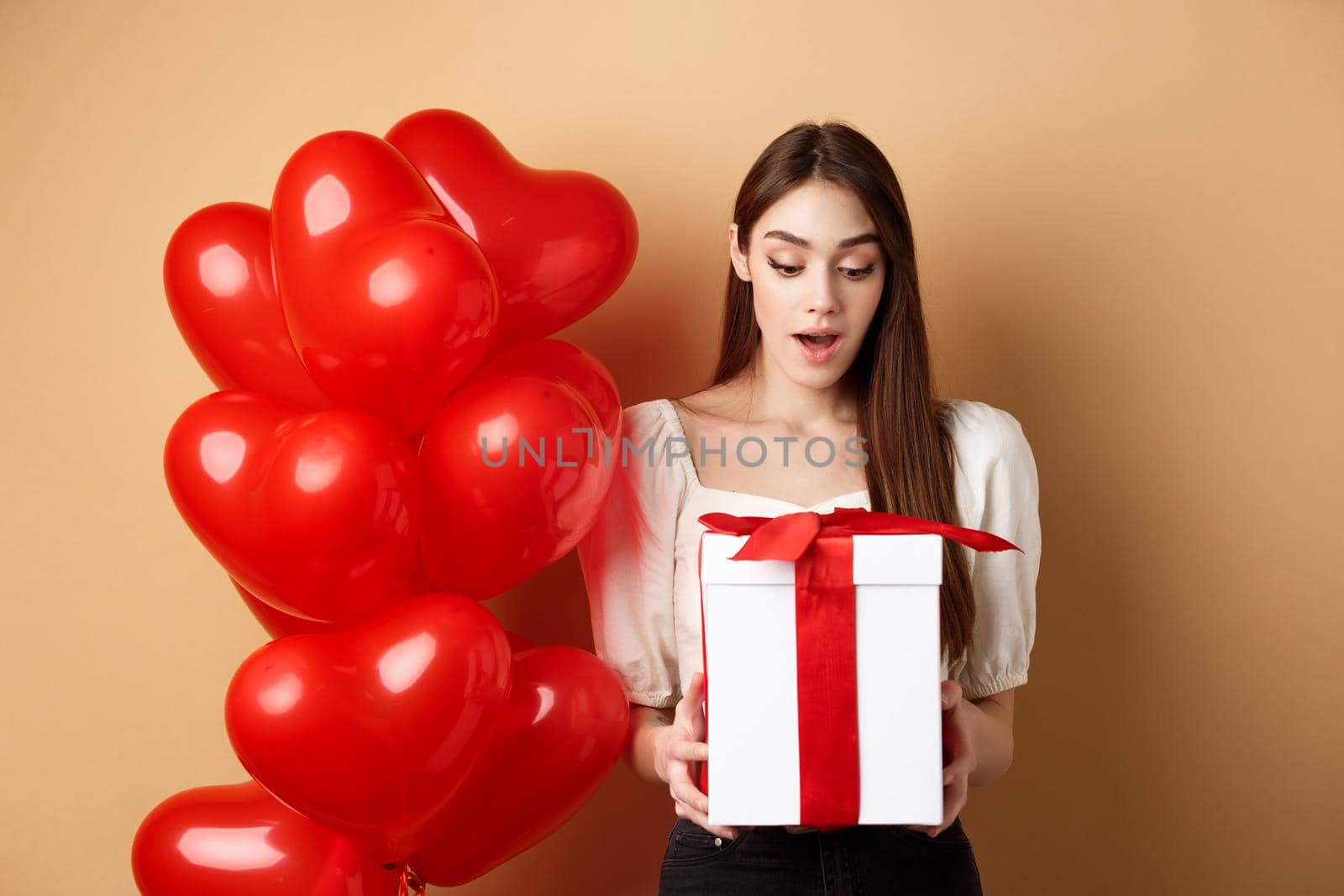 Surprised beautiful woman in romantic outfit, standing near heart balloons and looking at her gift on Valentines day, standing on beige background.