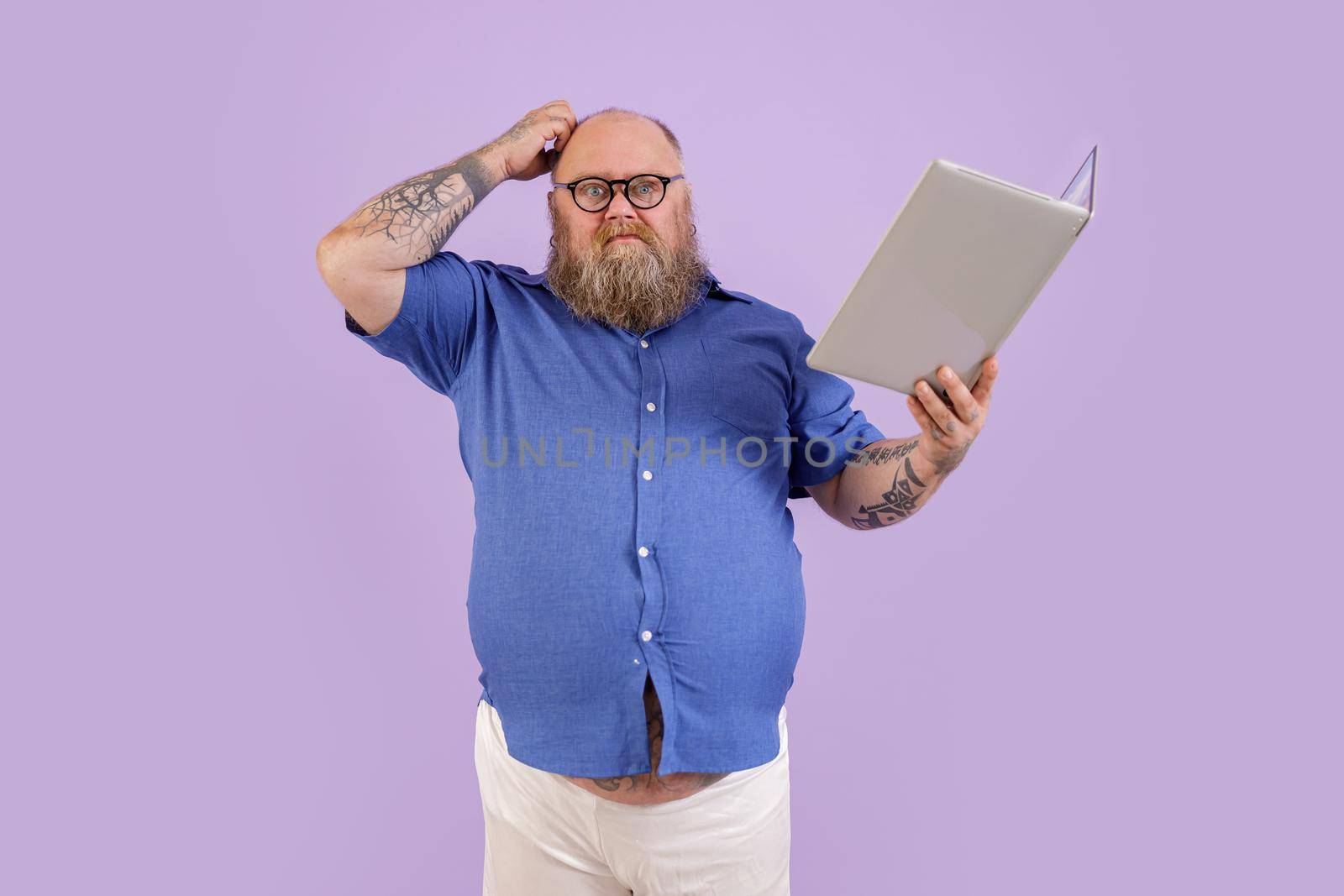 Doubting man with overweight shirt scratches head holding laptop as book on purple background by Yaroslav_astakhov
