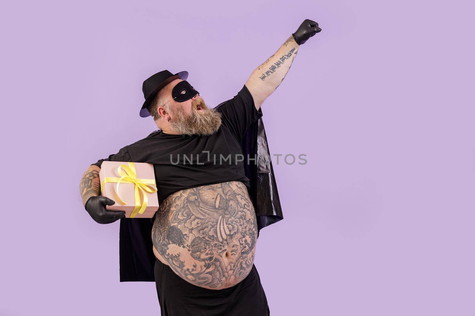 Funny gentleman with overweight wearing hero costume holds present on purple background by Yaroslav_astakhov