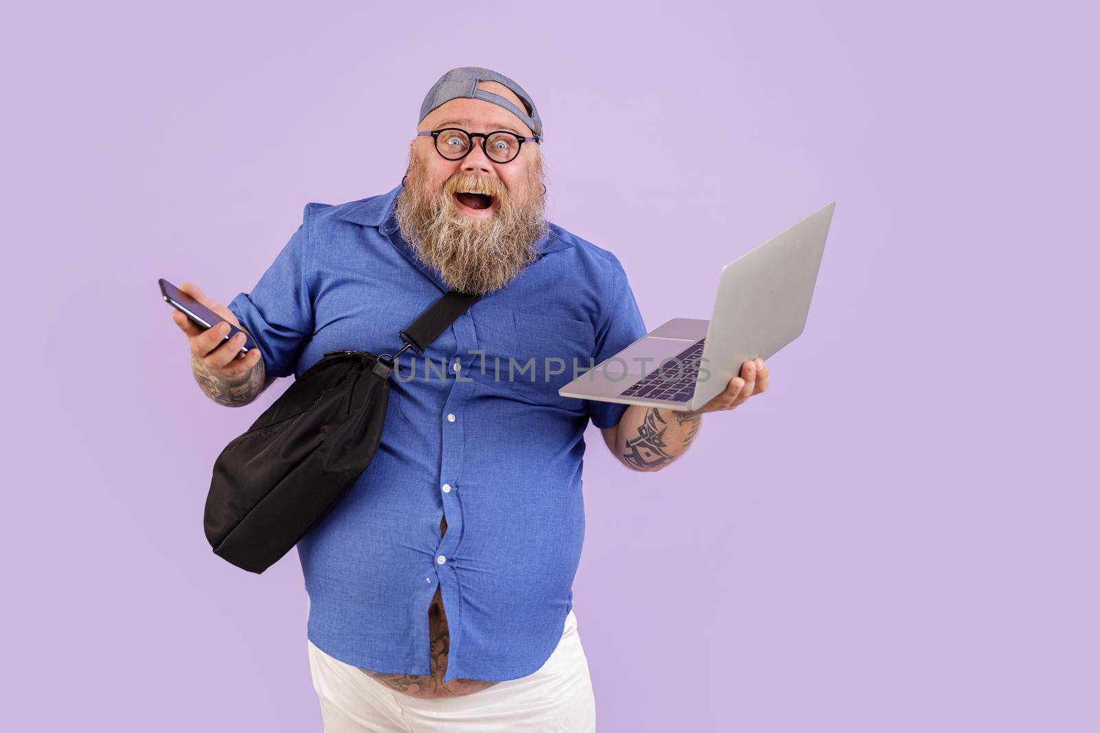 Expressive happy obese man with crossbody bag holds cellphone and contemporary open laptop standing on purple background in studio