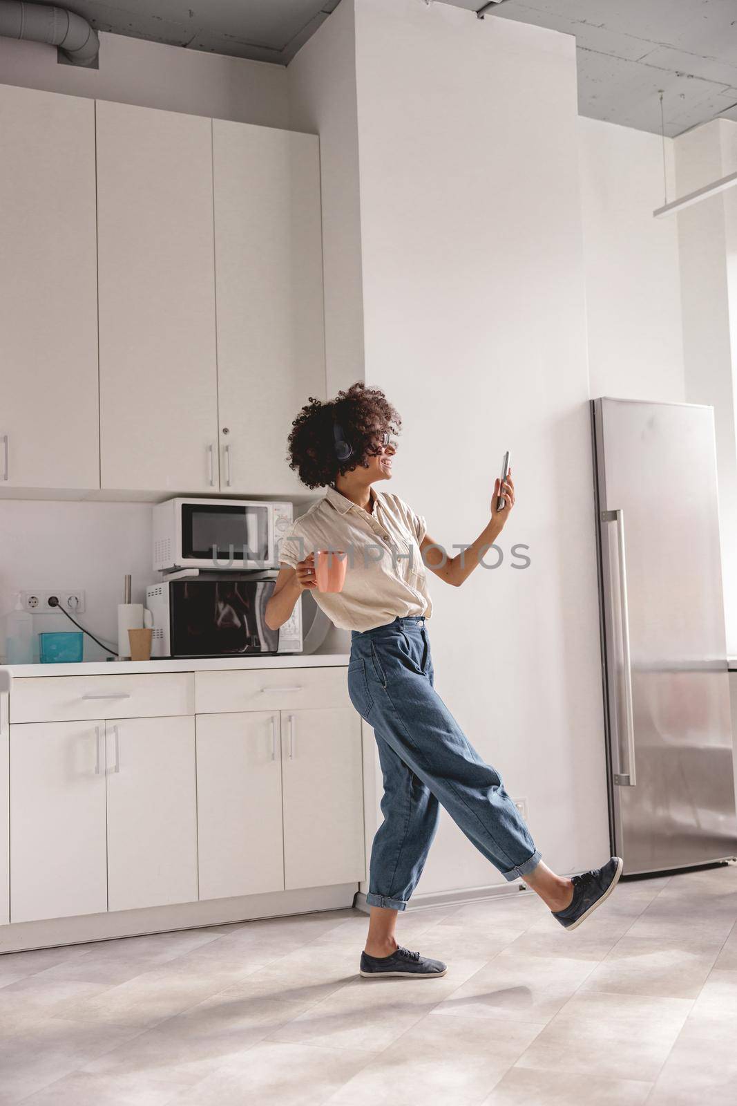 Full-length photo of happy pretty woman holding cup while dancing on the kitchen and using headphones