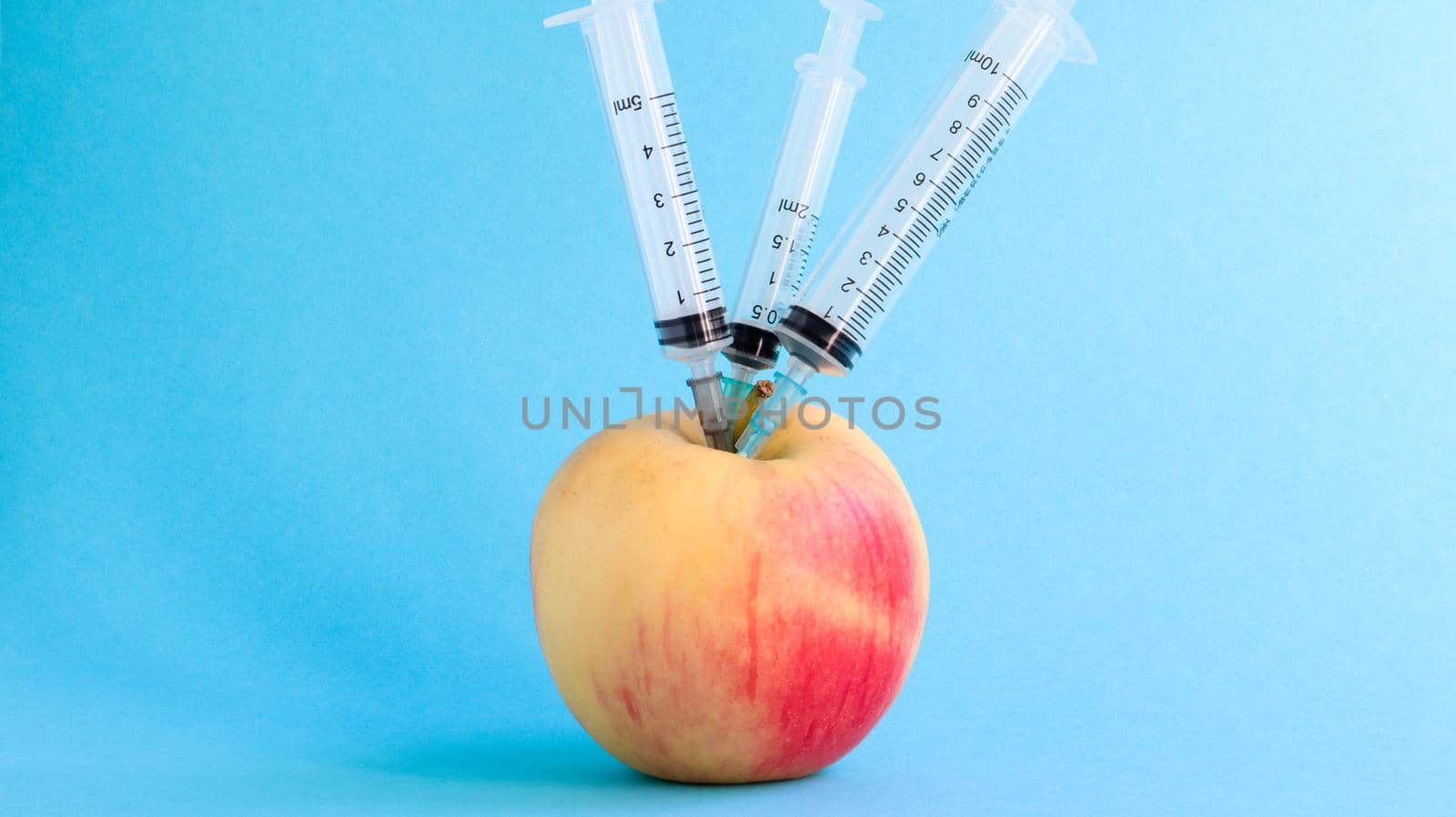 Three different medical syringes sticking deep into an apple on a blue background. The fruit is strewn with syringes. A ripe red apple pierced from all sides by syringes. Healthcare or cosmetology