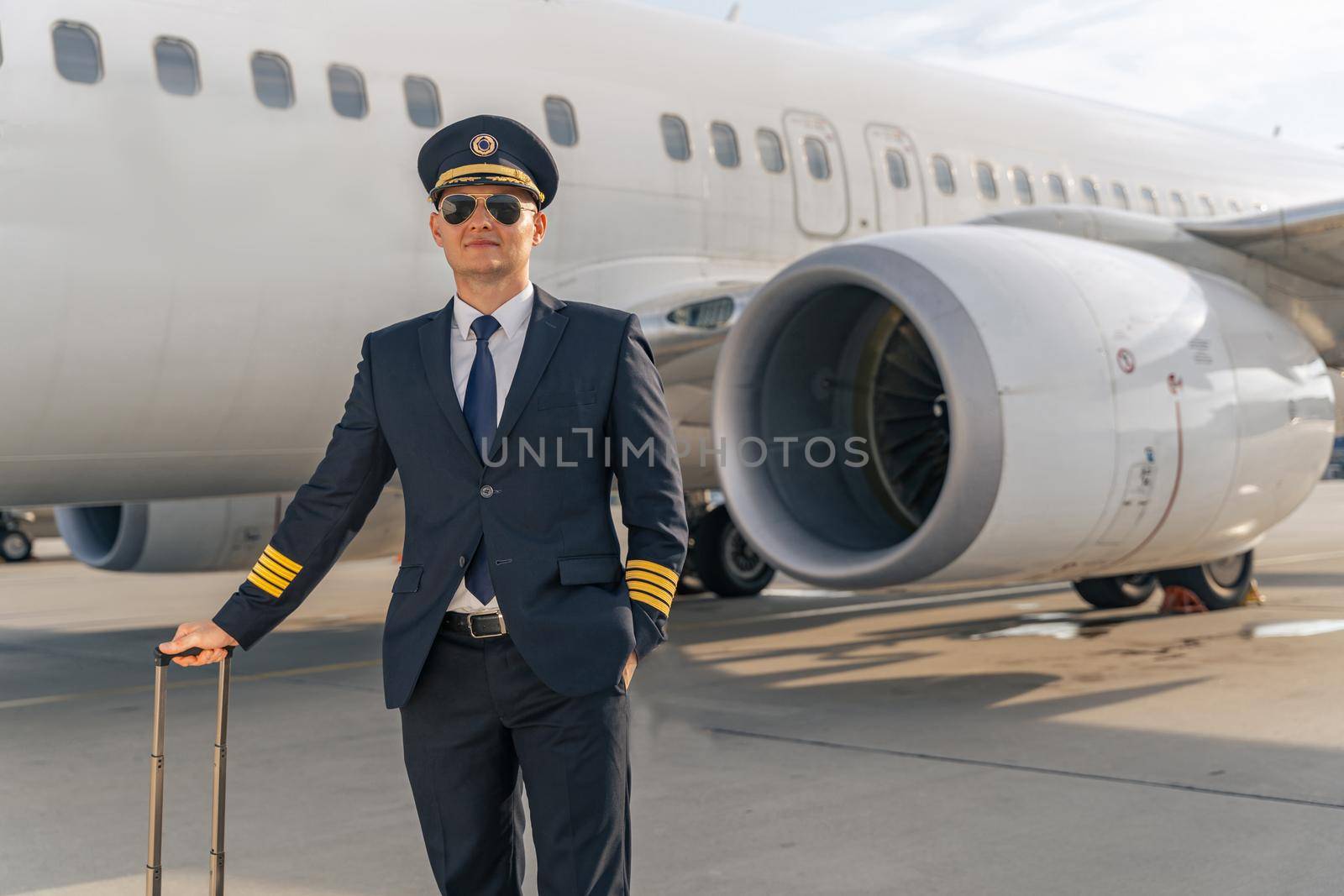 Stylish pilot in sunglasses holding travel bag with airplane on the background. Profession concept