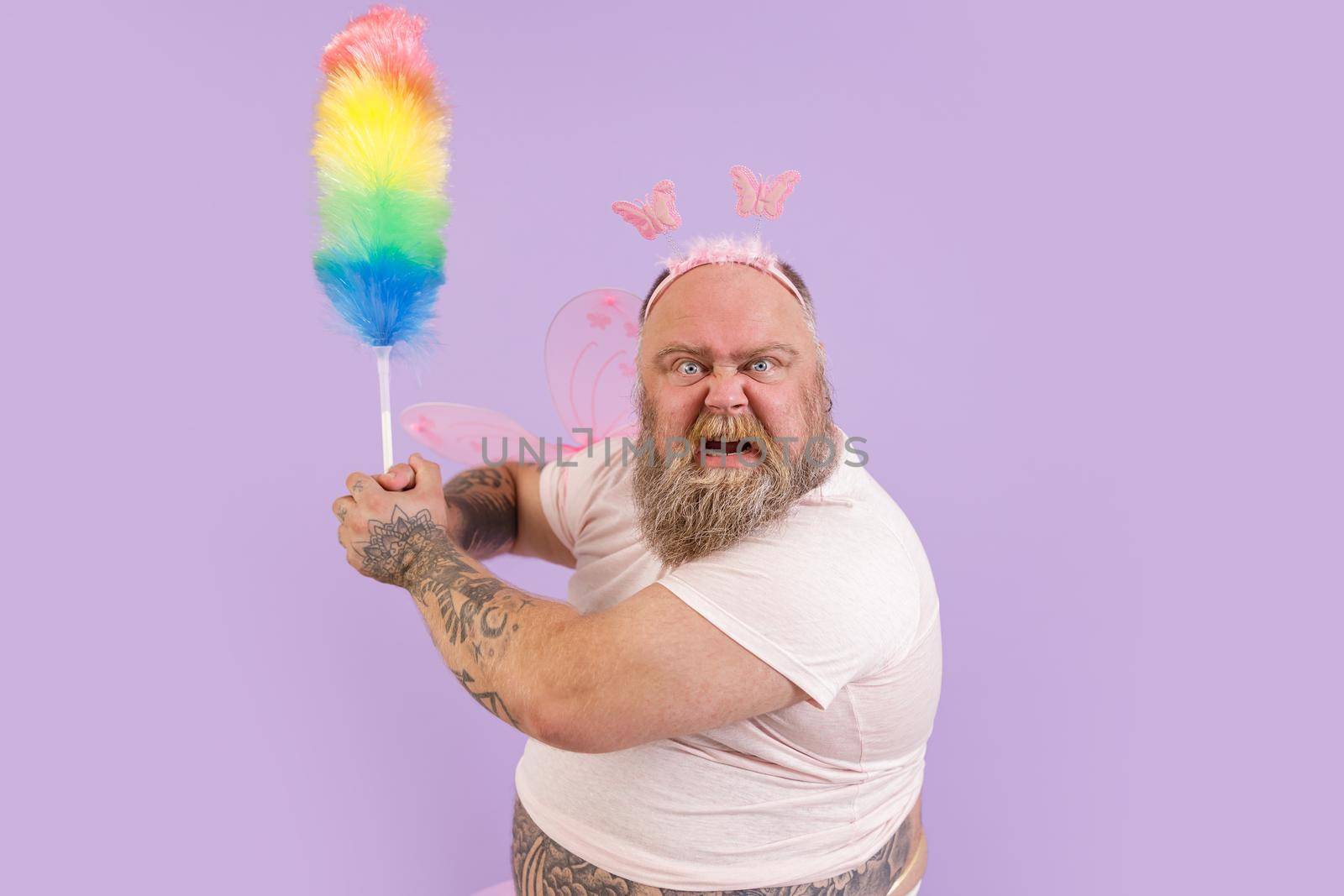Funny angry bearded plump man in fairy suit with wings attacks with fluffy dust brush on purple background in studio