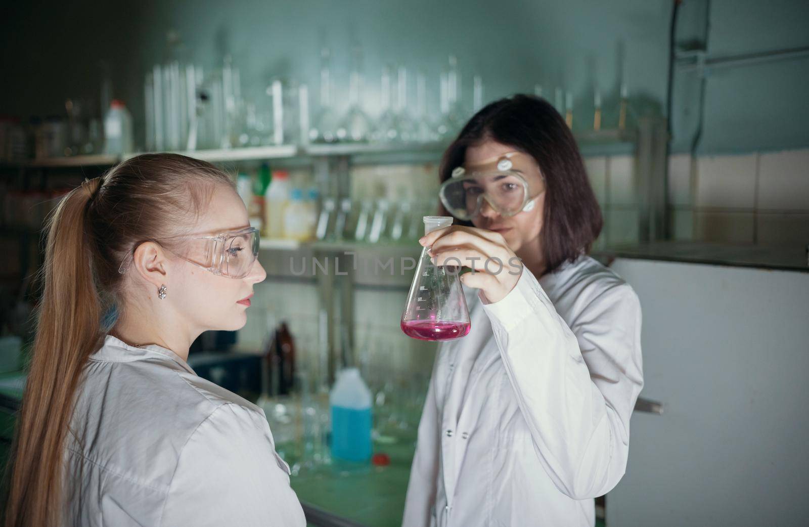 Two young woman in chemical laboratory holding a flask with pink liquid in it. Mid shot