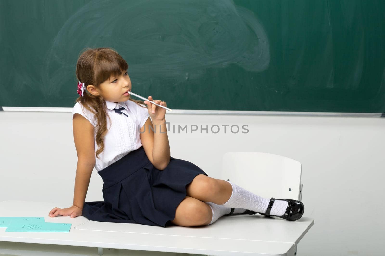Cute girl sitting on desk in classroom. Adorable girl student in white blouse and blue skirt holding pen in her hand posing at blackboard. School and education concept