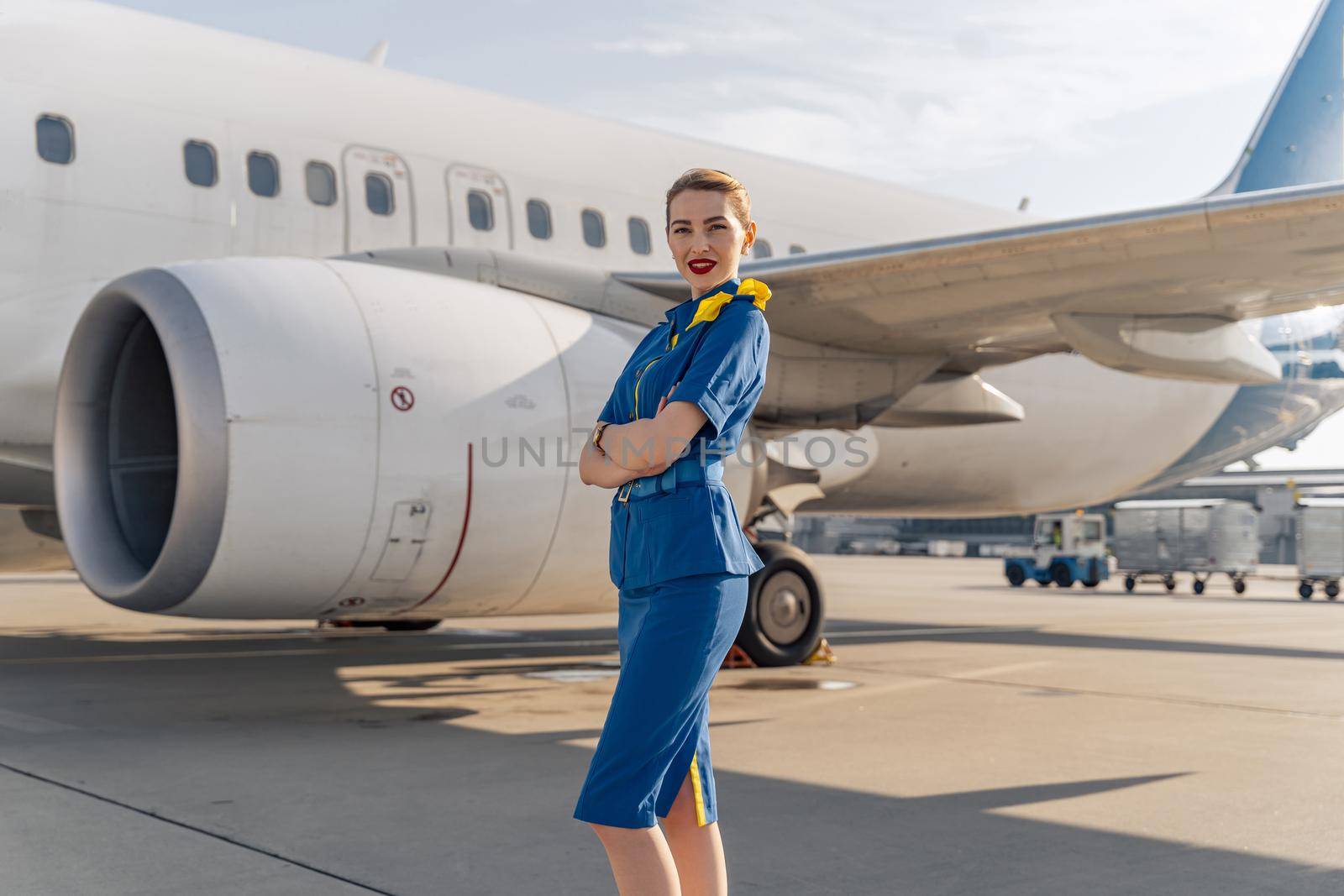 Happy beautiful stewardess crossed her arms while posing with white plane on the background