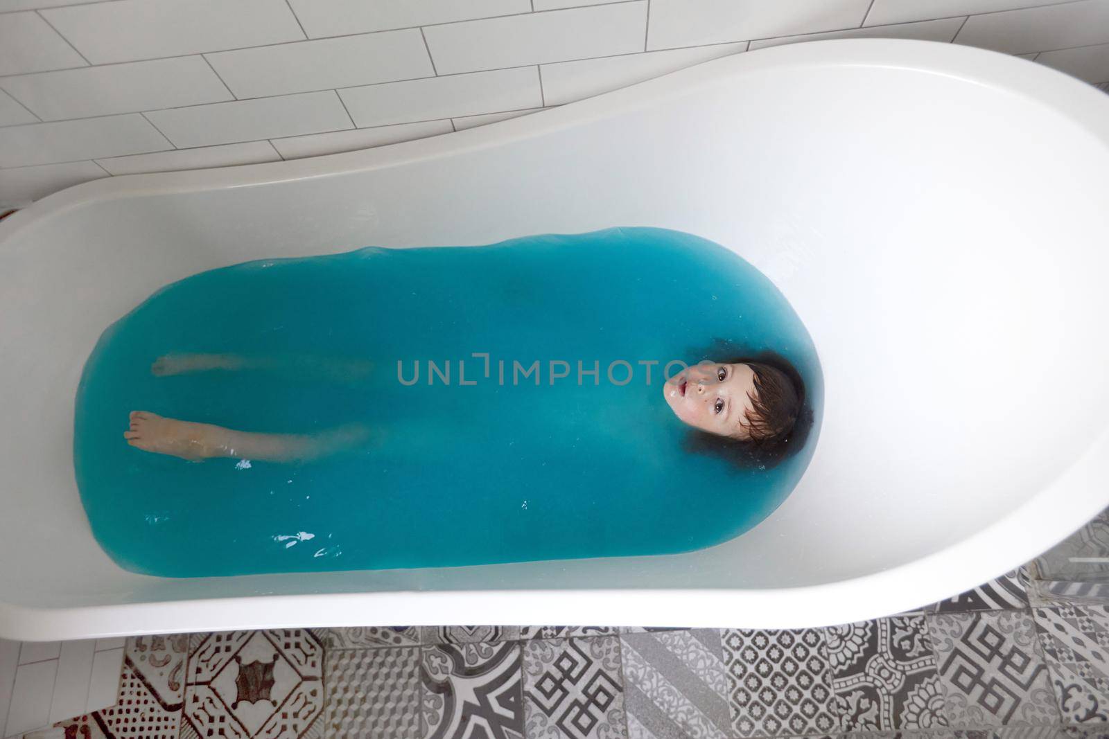 Baby boy swims on his back in blue water in bathtub. Top view of adorable little kid lying relaxing during hygiene routine