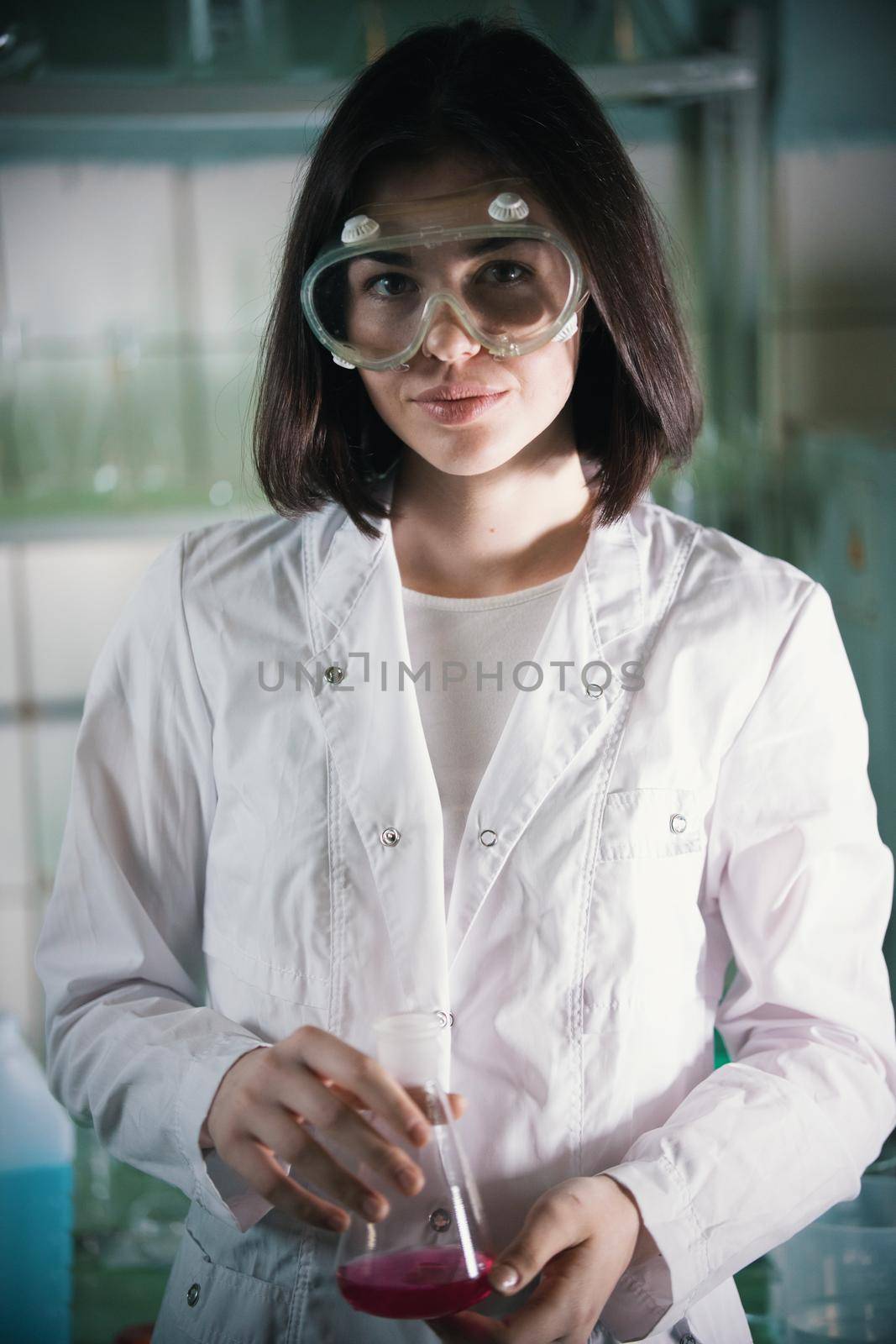 Chemical laboratory. Young woman in work glasses holding a flask and looking in the camera. Portrait