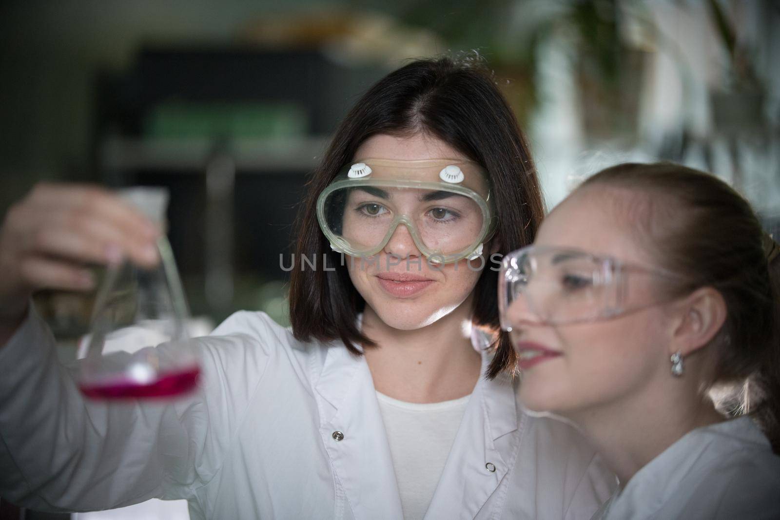 Chemical laboratory. Two young woman holding a flask with pink liquid in it, smiling. Close up