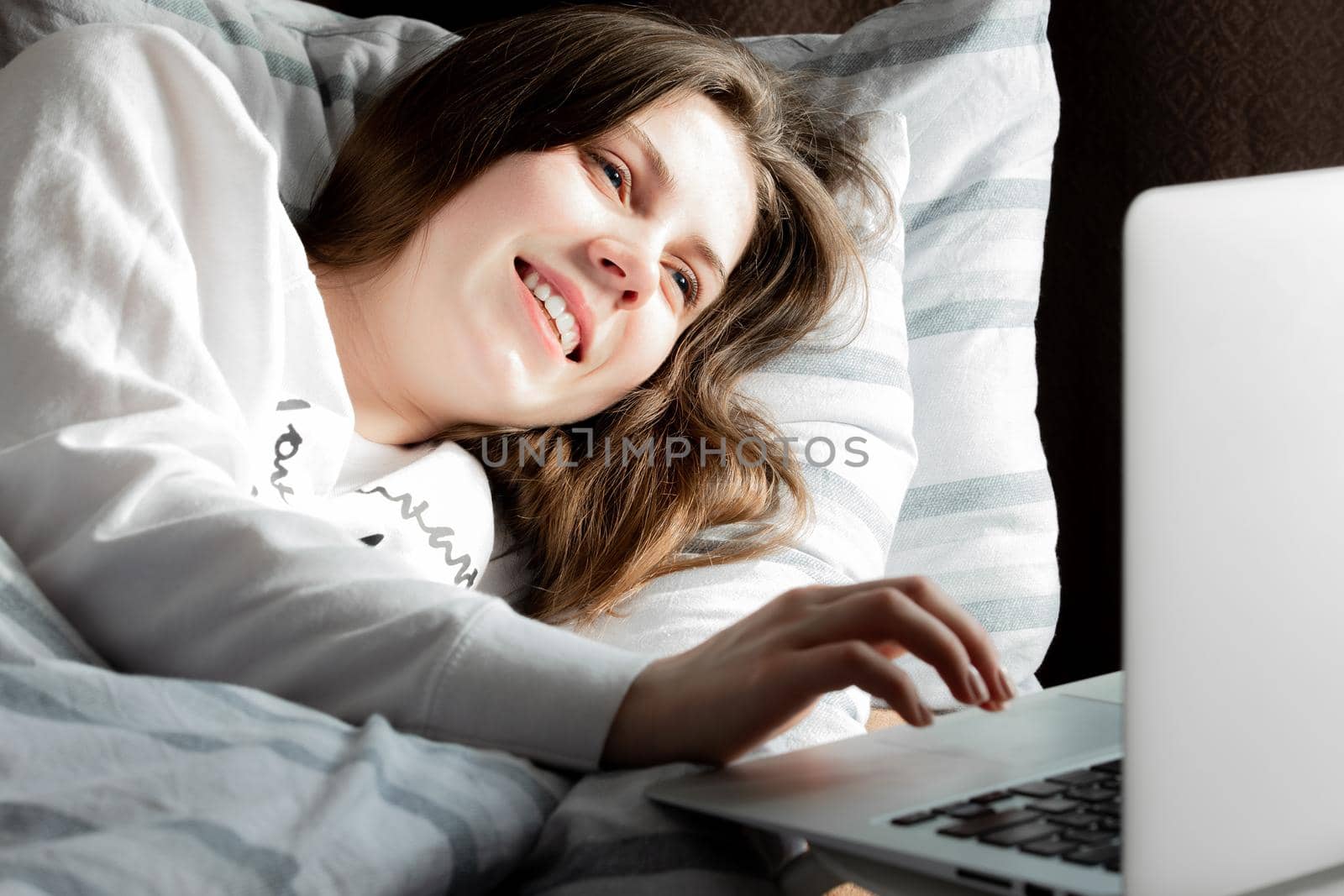 Young cute girl smiles on a sunny day in a white jacket lying on the bed and looking into a laptop.