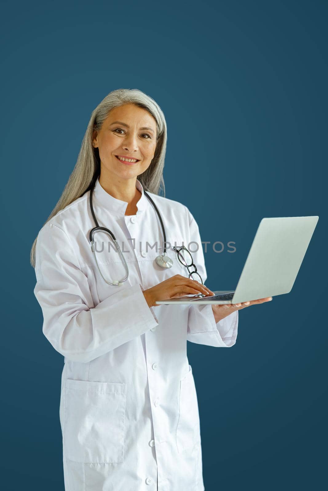 Cheerful mature lady doctor in white robe uses modern laptop standing on blue background in studio. Telemedicine patient service